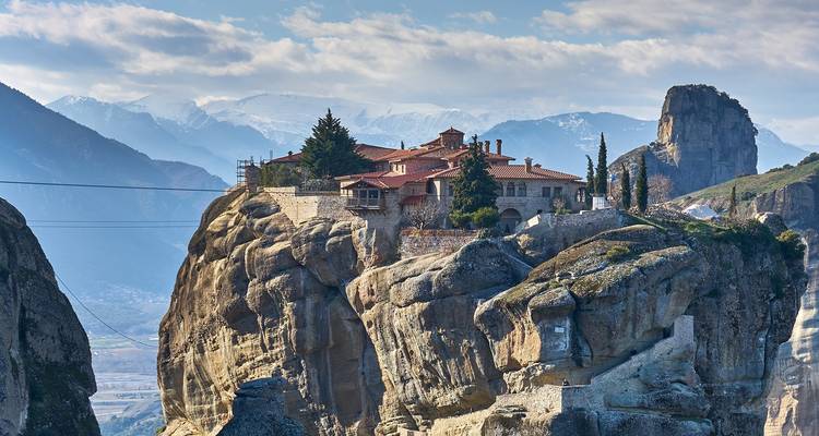Monastery perched on a large rock formation in Meteora, Greece, with mountains in the background.