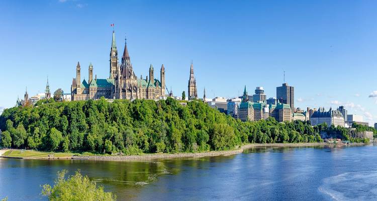 Parliament Hill’s Gothic towers rise above lush trees and the Ottawa River on a clear day.