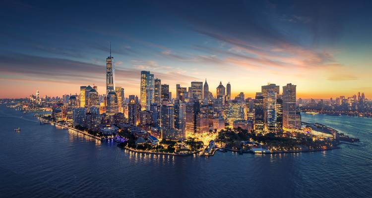 Lower Manhattan glows with city lights at twilight surrounded by the Hudson River.