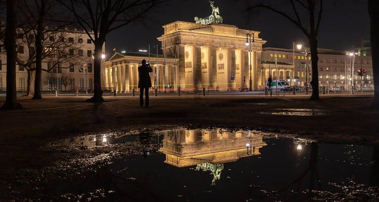 Porte de Brandebourg illuminée la nuit avec des traînées de lumière en pose longue et son reflet dans une flaque d'eau au premier plan.