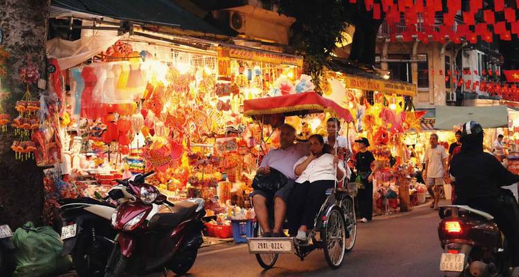 Scène de rue la nuit avec un cyclo transportant des gens devant des rangées de lanternes colorées.
