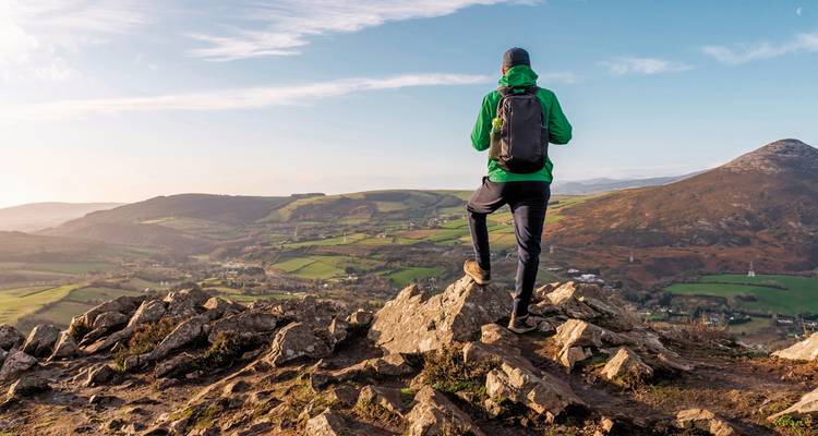 Randonneur sur un sommet rocheux avec vue sur des champs verts et des montagnes.