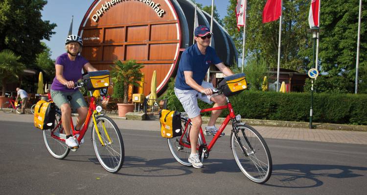 Deux personnes à vélo passant devant un grand tonneau de vin avec de la verdure et des drapeaux en arrière-plan.