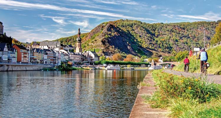 Deux cyclistes le long d'une promenade au bord de la rivière avec une ville historique et une église.