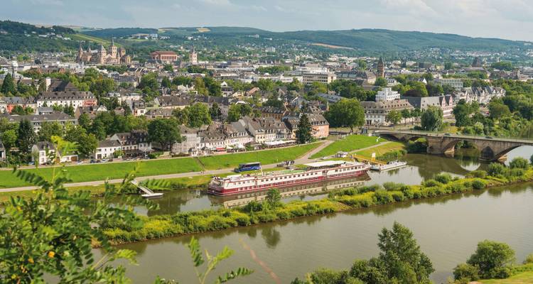 Vue panoramique d'une ville au bord de la rivière avec un bateau de croisière et une végétation luxuriante en arrière-plan.