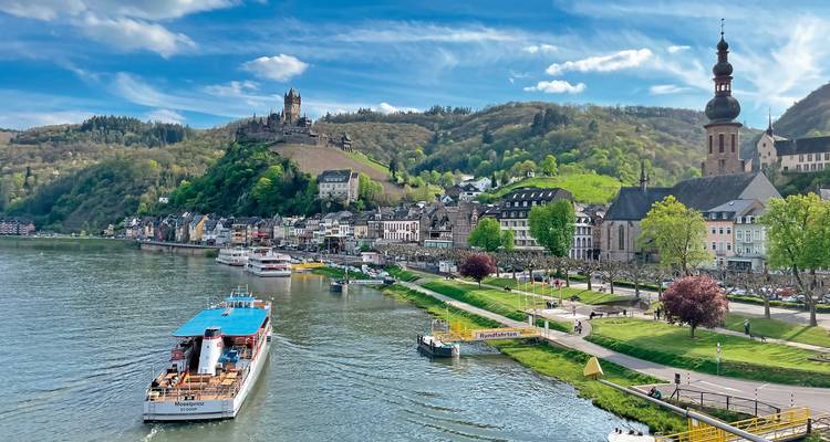 Un barco fluvial se acerca a un pueblo pintoresco con un castillo en una colina.