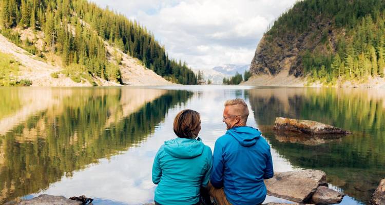 Deux personnes assises au bord d'un lac entouré d'arbres et de montagnes.