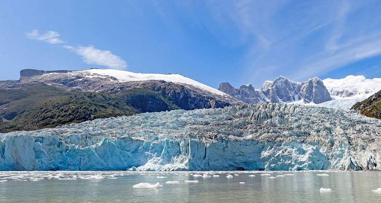 Dramatischer blauer Gletscher, der auf ruhiges Wasser trifft, mit schneebedeckten Bergen und blauem Himmel im Hintergrund.