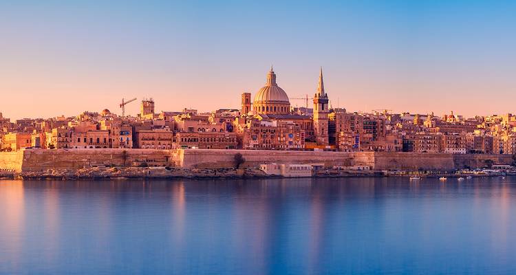 Panorama de la hora dorada de una ciudad mediterránea fortificada con catedral de cúpula reflejada en agua azul tranquila.