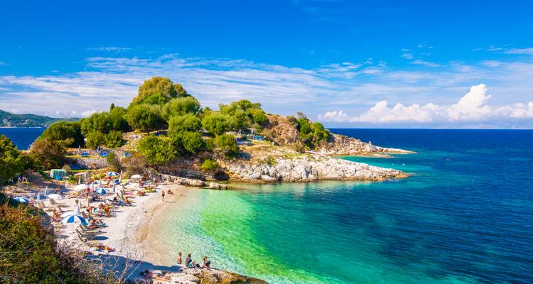Cala de playa soleada con agua turquesa, bañistas, sombrillas y vegetación rocosa en la costa jónica.