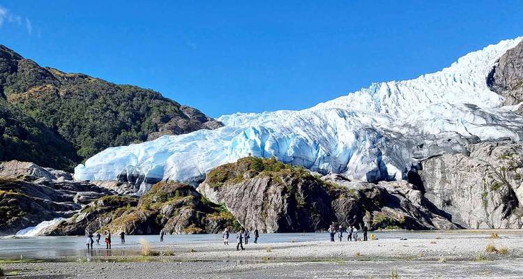 Des aventuriers marchent à travers une vaste plaine vers un glacier bleu éclatant sous un ciel dégagé.