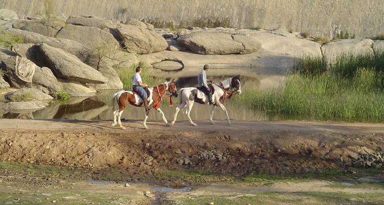 Twee mensen die paarden berijden naast een rivier met rotsen.
