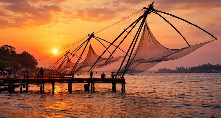 Silhouetted fishermen stand on a wooden jetty with giant Chinese fishing nets against a fiery sunset over the sea.