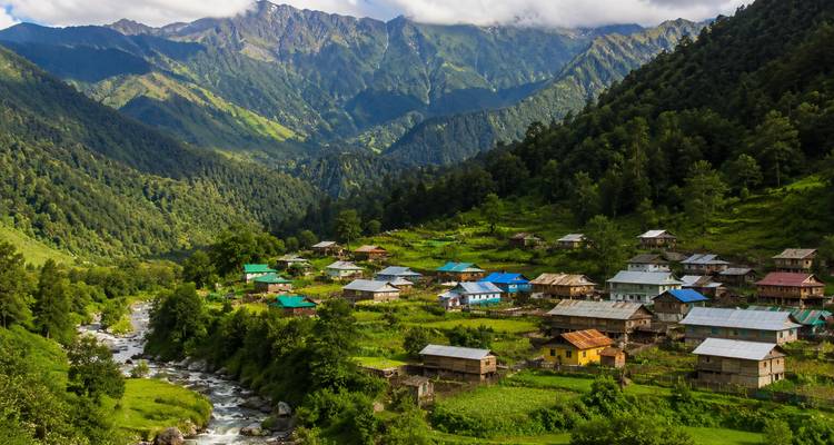 Mountain valley dotted with colorful village rooftops beside a rushing river and forested slopes.