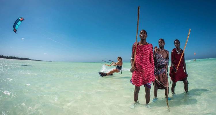 Personas con vestimenta tradicional con un kitesurfista en aguas poco profundas.
