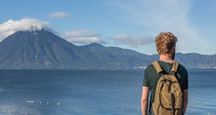 Homme aux cheveux roux avec un sac à dos debout près d'un lac avec des montagnes en arrière-plan.