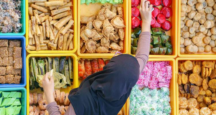 Vista aérea de un vendedor de mercado seleccionando bocadillos malayos coloridos de bandejas cuidadosamente organizadas.
