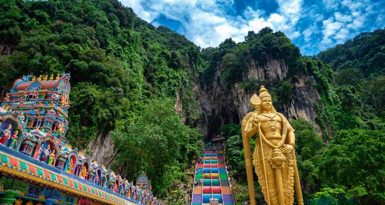 Façade colorée de temple hindou avec une imposante statue dorée de Murugan et un escalier arc-en-ciel sur fond de falaises calcaires couvertes de jungle.