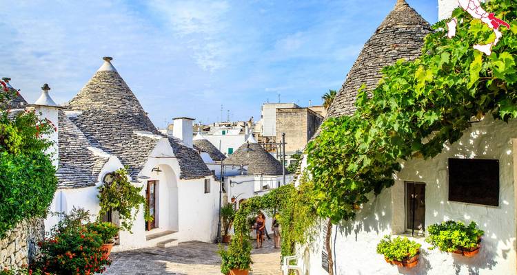 Casas trulli encaladas con techos cónicos de piedra bordean un sendero estrecho lleno de vegetación en Alberobello.