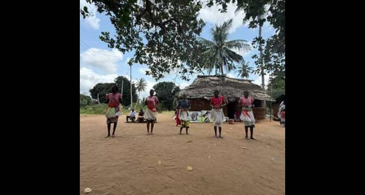 Grupo de personas con atuendo tradicional paradas en un área arenosa con árboles.