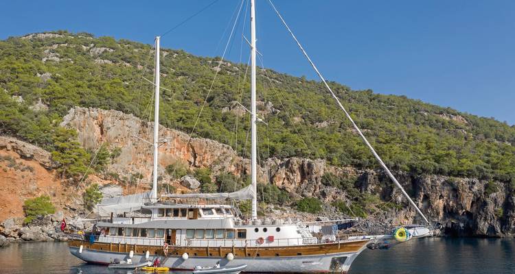 Zeilboot geankerd bij een rotsachtige kustlijn met bomen en een heldere lucht.