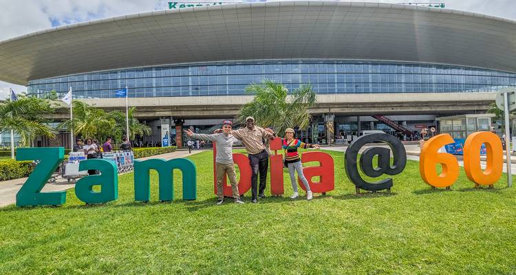 Touristes posant avec un panneau Zambie@60 devant un bâtiment moderne.