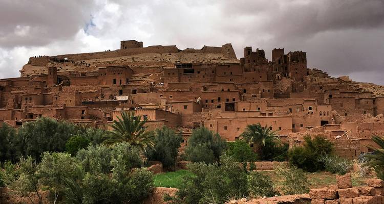 Antiguo ksar de ladera con edificaciones y torres de tierra que se alzan sobre una hilera de palmeras bajo un cielo melancólico lleno de nubes.