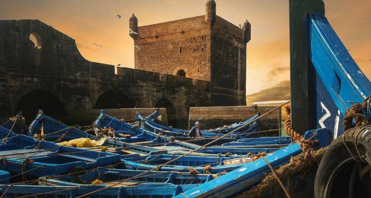 Fisherman sits among bright blue wooden boats with Essaouira’s harbour fort glowing in golden sunset light.