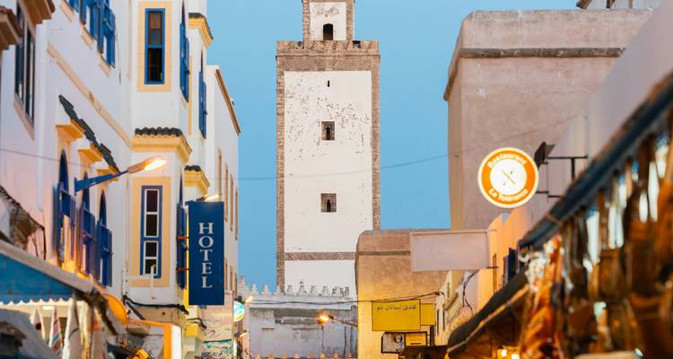 Evening scene of a white minaret rising above bustling market stalls and neon signs in Essaouira.