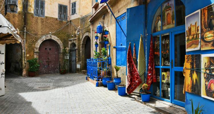 Quiet cobblestone courtyard with blue doors, pots of plants, and colorful rugs displayed outside a shop.