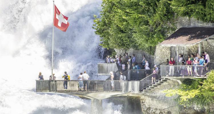 Des gens sur une plateforme près d'une grande cascade avec le drapeau suisse.