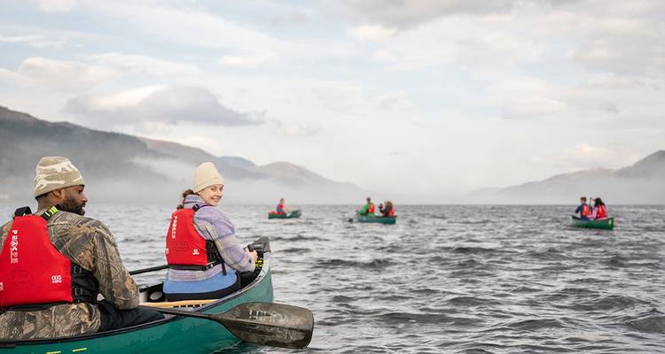 Mehrere Kanus gleiten über einen nebligen schottischen Loch, umgeben von Bergen, während Paddler das Wasser genießen.