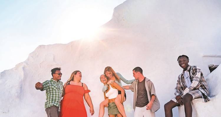 Gruppe von Freunden, die im hellen Sonnenlicht vor der weiß getünchten kykladischen Architektur auf Mykonos lachen.