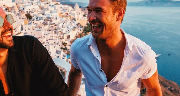 Twee vrienden lachen samen boven de caldera van Santorini met witgekalkte gebouwen op de achtergrond.