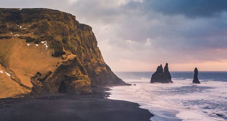 Stimmungsvoller Sonnenuntergang über dem schwarzen Sandstrand von Reynisfjara mit dramatischen Basalt-Felstürmen, die aus dem Atlantik herausragen.