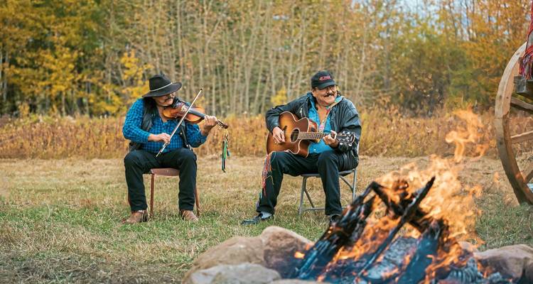 Zwei Männer spielen Musikinstrumente in der Nähe eines Lagerfeuers auf einer grasbewachsenen Wiese mit herbstlichem Laub.