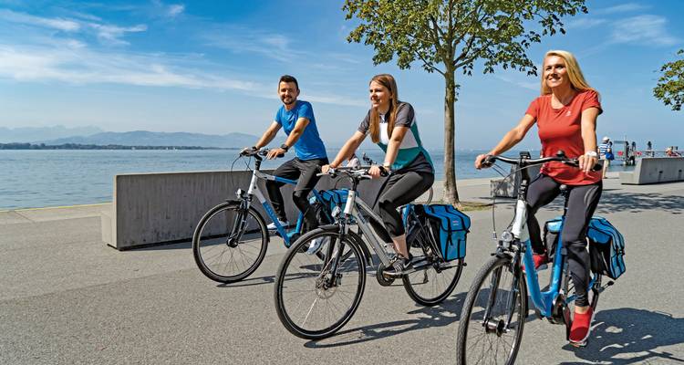 Tres personas andando en bicicleta por un sendero junto al lago con montañas visibles.
