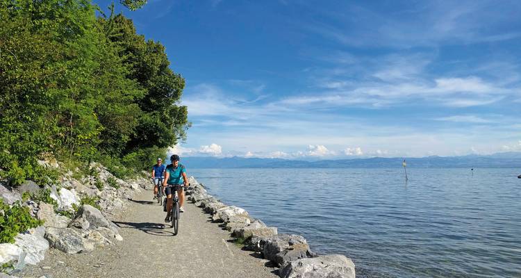 Ciclistas pedaleando por un sendero junto al lago con montañas al fondo.