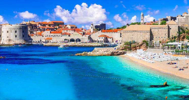 Historic walled city of Dubrovnik with bright turquoise Adriatic waters and sandy beach in foreground.