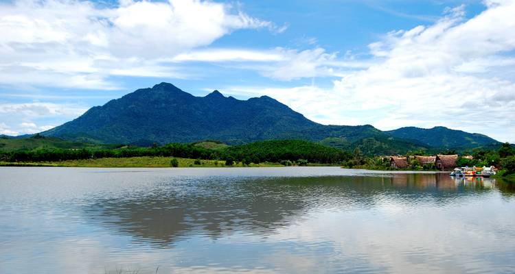 Lago tranquilo reflejando una montaña boscosa bajo cielos brillantes del mediodía