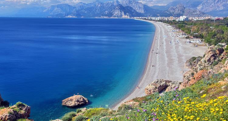 Pebbly beach with mountains and city in the background.