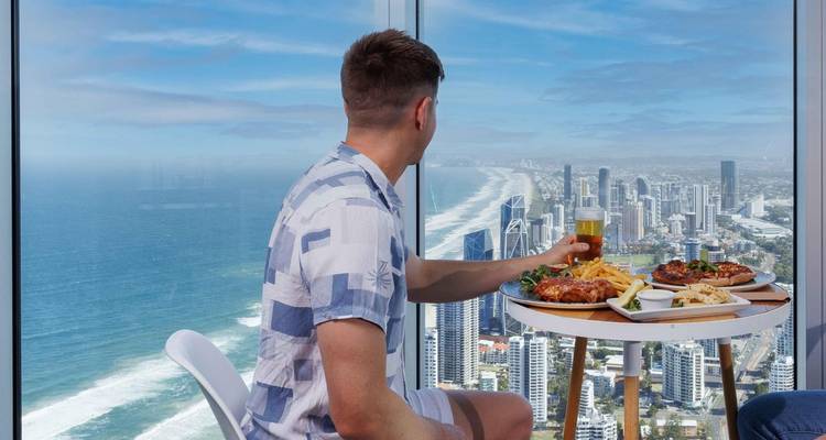 Una persona disfrutando de una comida con vista a una ciudad costera desde un rascacielos.