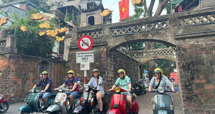 Un grupo de personas en scooters se detuvo en una intersección de calles cerca de un arco histórico y edificios.