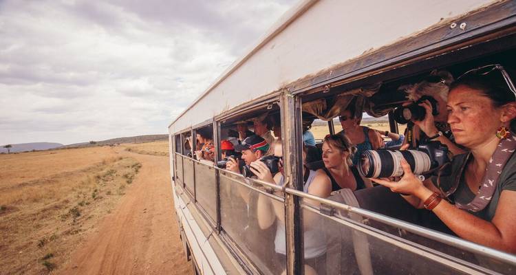 Groupe de touristes prenant des photographies depuis un bus de safari sur une route de terre.