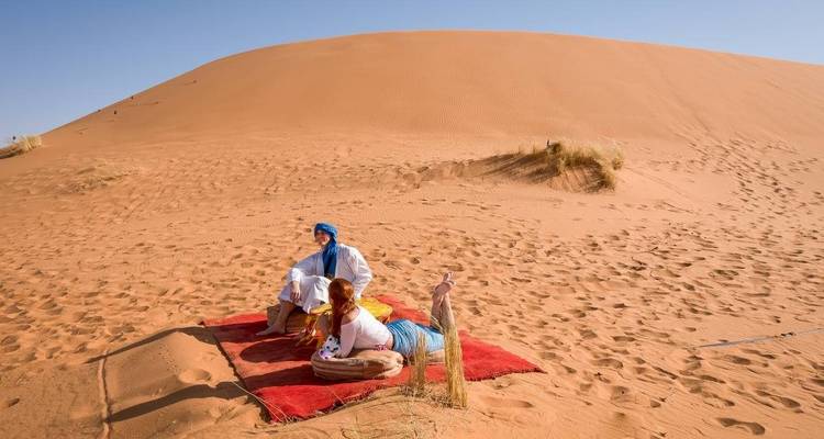 Una pareja relajándose en una alfombra roja al pie de una enorme duna de arena bajo los cielos desérticos despejados.