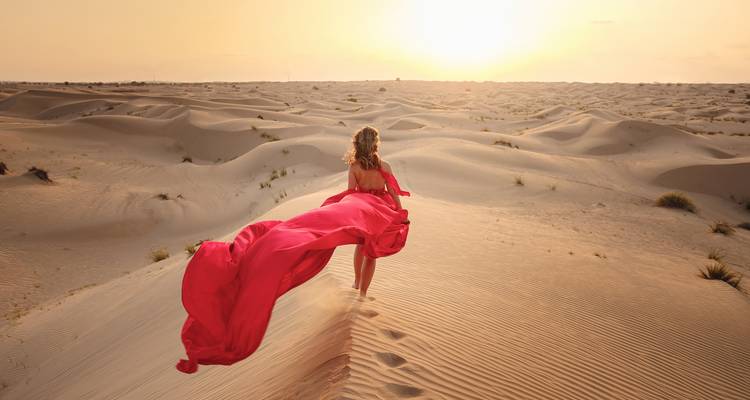 Woman in a flowing red dress running through desert sand dunes.