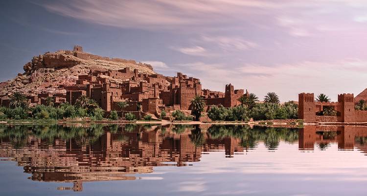 The mud-brick citadel of Ait Benhaddou is reflected perfectly in tranquil water beneath a pastel sky.