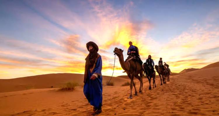 A blue-robed guide leads a camel caravan across glowing dunes beneath dramatic sunset clouds.