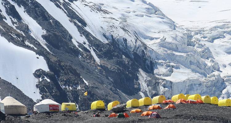 Camp de base avec des tentes jaunes et des yourtes au pied d'un glacier, avec des montagnes en arrière-plan.