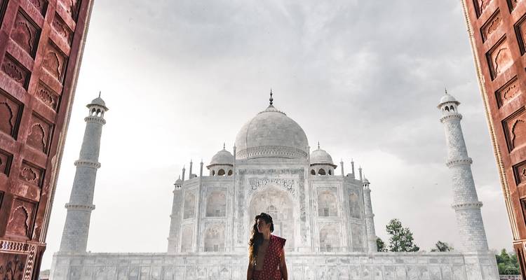 Une femme vêtue d'une tenue rouge traditionnelle contemple le Taj Mahal en marbre encadré par la porte ornementée en grès rouge sur un jour nuageux.